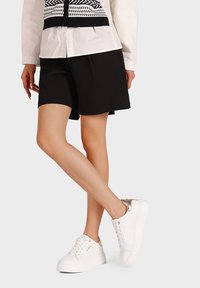 Sneakers bianchi con una finitura testurizzata e dettagli dorati, abbinati a pantaloni corti neri e una camicia bianca con un motivo geometrico nero.