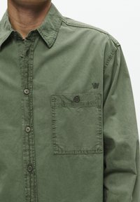 Camicetta verde oliva con apertura frontale a bottoni, taschino sul petto e delicati dettagli di cucitura diagonale. Realizzata in un tessuto leggero e morbido.