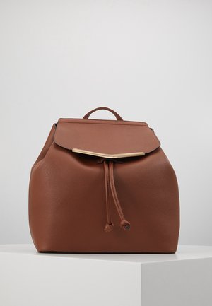 Mochila de cuero marrón con forma redondeada, acentos de hardware dorado, cierre de cordón y tapa con solapa, presentando una superficie texturizada.