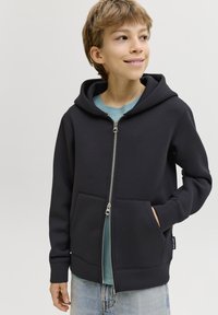 Sweat à capuche noir zippé en mélange de coton, doté d'une capuche, de poches avant et d'une fermeture éclair argentée. Porté par-dessus un t-shirt bleu clair et un jean en denim.