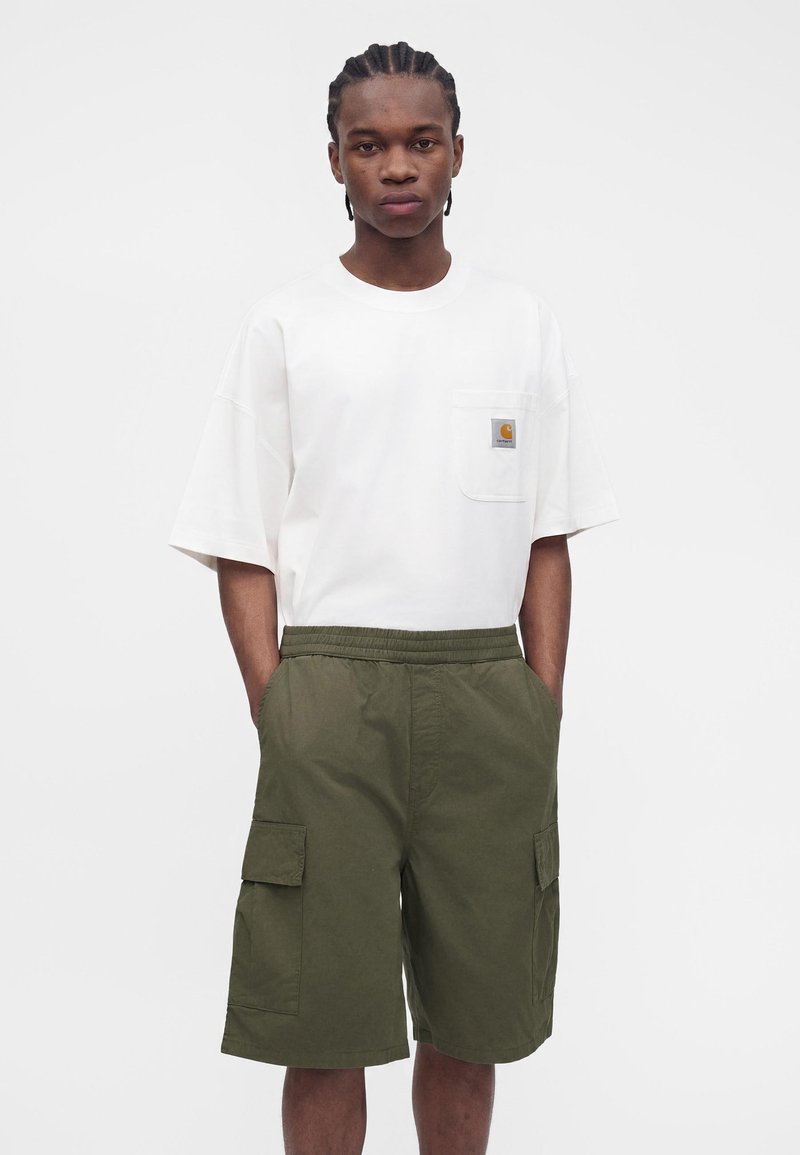 Mann steht mit den Händen in den Taschen, trägt ein weißes Kurzarmhemd mit Tasche und olivgrüne Cargo-Shorts.