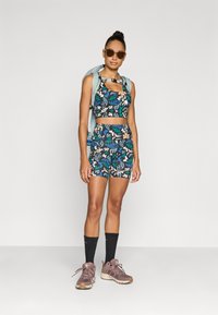 Σετ αθλητικού σουτιέν και σορτς με λουλουδάτο σχέδιο σε σκούρα χρώματα. Φορεμένο με γκρι σακίδιο, μαύρες κάλτσες μέχρι το γόνατο και γκρι-καφέ παπούτσια πεζοπορίας.