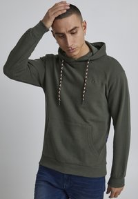 Olive green hoodie with a front pocket, featuring a textured fabric and patterned drawstrings. Includes logo detailing on the chest.