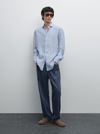 Camicia a righe blu chiaro, taglio rilassato, abbinata a pantaloni in denim scuro e scarpe slip-on in suede marrone. Modello che indossa occhiali da sole neri.