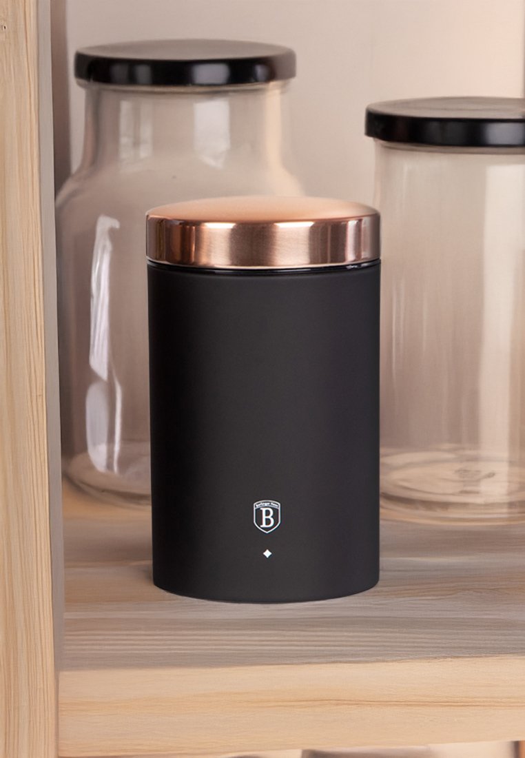 Contenitore di stoccaggio nero opaco con coperchio in rame, presenta un logo e un accento a forma di diamante alla base, posizionato su uno scaffale di legno vicino a barattoli di vetro.
