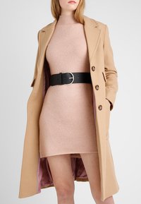 Un manteau en camel avec des revers en pointe sur une robe rose ajustée. Le manteau a des boutons marron et présente une ceinture noire avec une boucle circulaire en argent.