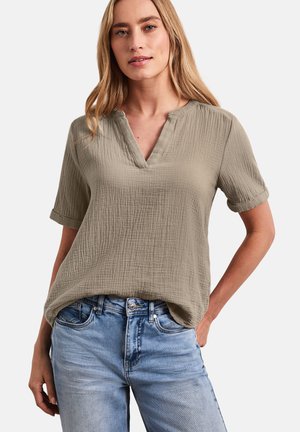 Vrouw draagt een beige blouse met korte mouwen en een gespleten hals, gemaakt van een textuurstof, gecombineerd met lichtblauwe spijkerbroek, tegen een witte achtergrond.