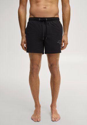 Mann trägt schwarze Badeshorts mit Kordelzug an der Taille und subtil dunklem Design am linken Bein, steht barfuß auf einfachem Hintergrund.