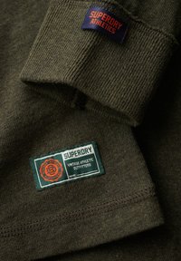 Grøn ribbet bomulds langærmet trøje med et marineblåt "Superdry Athletics" mærke og et rektangulært grønt label med orange detaljer.