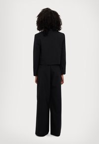 Blazer noir court à coupe ajustée, avec des manches longues et une texture lisse, associé à un pantalon noir à jambes larges.