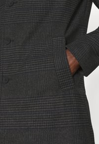 Ternet uldfrakke i mørkegrå og marineblå med tekstureret stof, der har sorte knapper og sidelommer. Skræddersyet pasform med strukturerede ærmer.