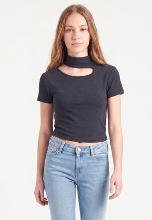 Jeune femme aux longs cheveux portant un crop top noir à manches courtes avec une découpe près du cou et un jean bleu clair, debout devant un fond uni.