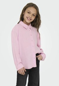 Chemise rose pâle à boutonner avec un col, des manches longues et une coupe décontractée, agrémentée de boutons roses et d'un tissu texturé. Jean noir porté.