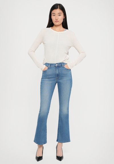 Lichtbeige lange mouwen top met een ronde hals, gecombineerd met mid-rise uitlopende blauwe jeans. Model draagt zwarte puntige hakken.
