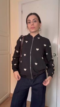 Femme portant une veste noire à fermeture éclair avec des motifs de cœurs blancs, un pantalon large et sombre, et un sac à épaule, debout à l'intérieur devant une porte.