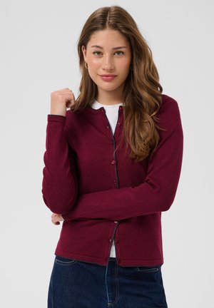 Jeune femme aux longs cheveux bruns portant un cardigan bordeaux boutonné sur un haut blanc et un jean bleu, posant les bras croisés.