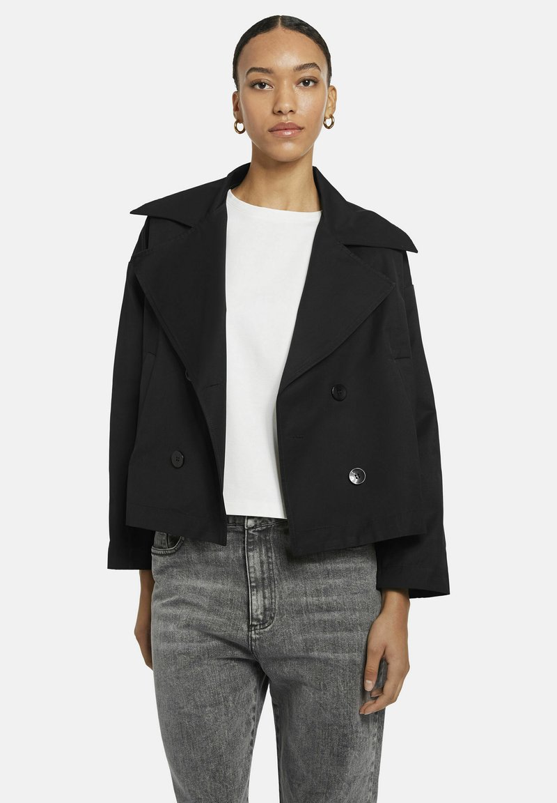 Vrouw in een zwart cropped dubbelzijdig jasje over een witte top en grijze hooggesneden jeans, staand tegen een witte achtergrond.