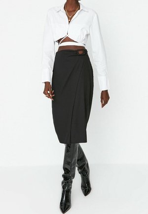 Femme portant une chemise courte blanche, une jupe portefeuille noire avec détail de boucle, et des bottes noires texturées montantes jusqu'aux genoux, debout sur un fond blanc.