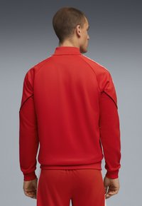 Mandlig model iført en rød atletisk træningsdragt, set fra ryggen, med en simpel grå baggrund, der fremhæver outfitets detaljer.