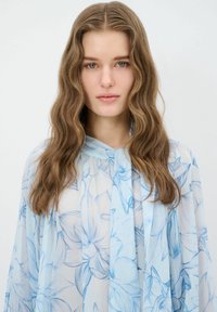 Lys blå blomstret bluse lavet af gennemsigtig stof. Har et flydende design med en bundet hals og bløde, bølgetexturerede ærmer.
