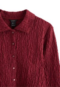 Bordeaux-farvet tekstureret skjorte med krave, der har subtile plisseringer og runde knapper foran. Brandlabel synlig indeni.