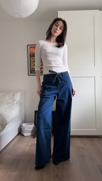 Fiatal nő áll egy hálószobában, fehér hosszú ujjú felsőt és derékban megkötött, magas derekú, bő szárú kék farmert visel.