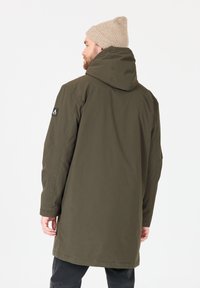Parka verde oliva con cappuccio, caratterizzata da una toppa con logo sul braccio superiore, taglio dritto e texture liscia. Indossata con un berretto lavorato a maglia beige.