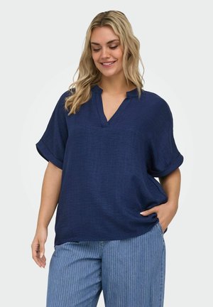 Lachende vrouw draagt een los vallend marineblauw V-hals T-shirt met korte mouwen en blauwe broek met fijne strepen, één hand in de zak, tegen een effen achtergrond.
