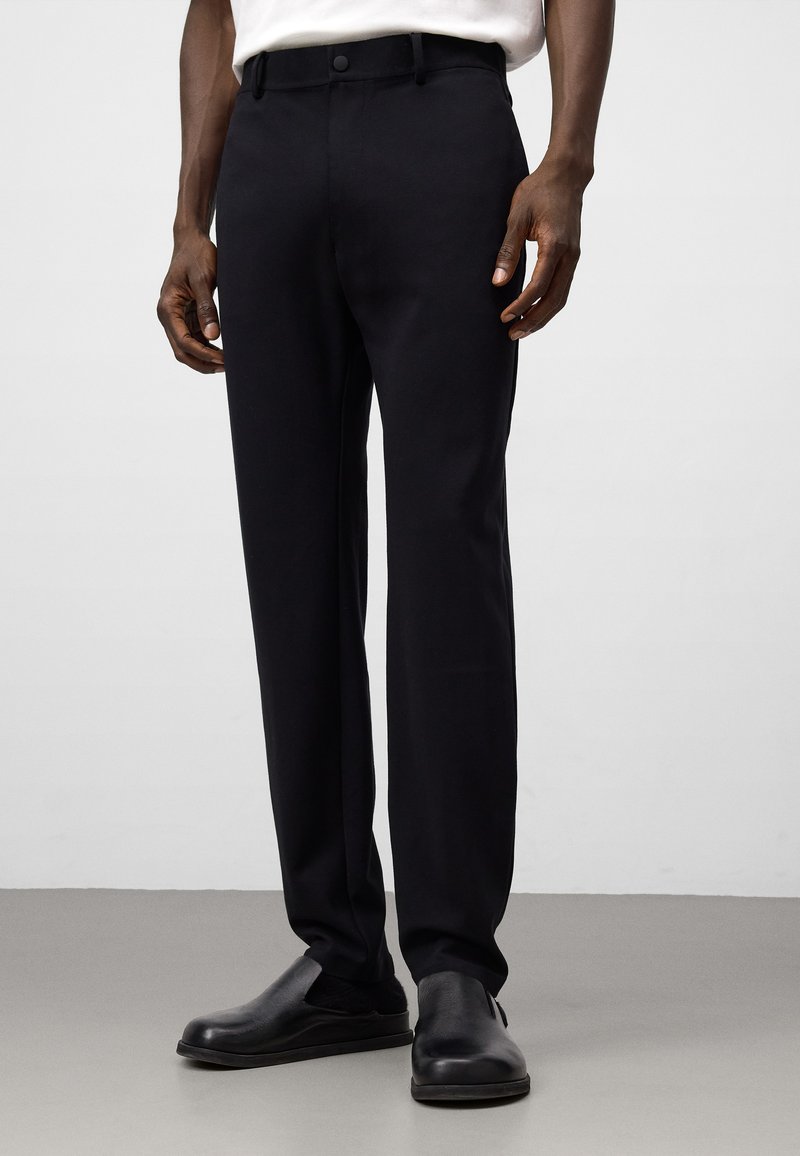 Pantaloni neri con una texture liscia, taglio dritto e vita standard. Presentano una chiusura con un bottone e tasche laterali.