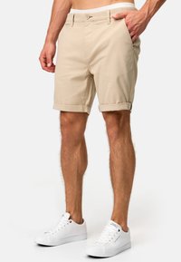 Beige bomulds-shorts med knaplukning og sidelommer, med rullede kanter. Båret med hvide sneakers.