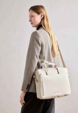 Sacoche beige pour ordinateur portable avec deux poignées et une bandoulière amovible. Dispose d'une poche zippée à l'avant et de côtés texturés. Portée par-dessus un blazer à carreaux.