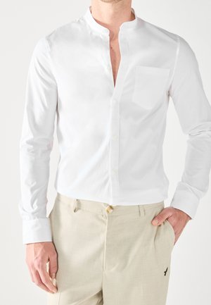 Mann trägt ein weißes Hemd mit Stehkragen und beige strukturierte Hose mit kleinem, besticktem Logo nahe der Tasche.