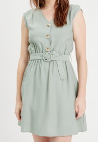 Robe vert clair sans manches avec un col en V, devant boutonné, taille cintrée avec une ceinture en tissu ornée d'une boucle ronde, texture lisse.