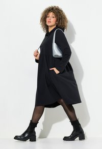 Schwarzes langärmeliges Kleid mit Kapuze, elastischem Bund und Seitentaschen. Kombiniert mit klobigen schwarzen Stiefeln und einer silbernen Handtasche. Jüngste Struktur.