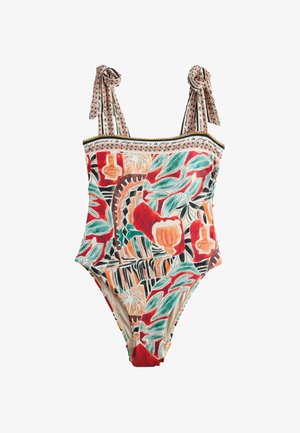 Femme debout pieds nus contre un mur beige, portant un maillot de bain coloré à motifs avec une jupe sarong nouée.
