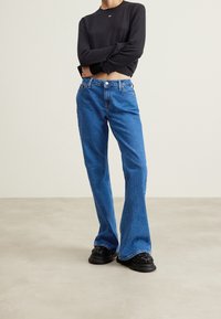 Blue denim flared jeans with a high waist, button closure, and five pockets, paired with a black long-sleeve top. Black textured shoes.