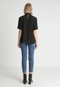 Svart blus med prickar och korta ärmar, med en nyckelhålöppning bak, kombinerad med blå jeans och svarta ankelstövlar. En plain bakgrund.