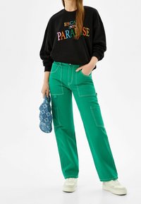 Person som bär en svart sweatshirt med färggfull texten "Escape Into Paradise", gröna cargobyxor, vita sneakers och håller en blå mönstrad hatt.