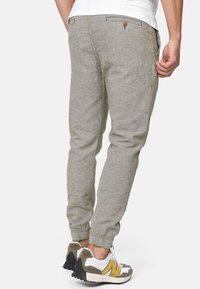 Homme portant un pantalon de jogging gris clair et des baskets blanches avec des accents jaunes, vu de dos sur un fond uni.