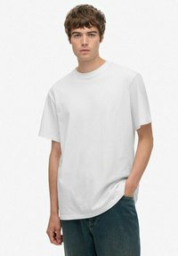 Unge mand med kort brunt hår, iført en ensfarvet hvid T-shirt med korte ærmer og blå jeans, stående med den ene hånd i lommen.
