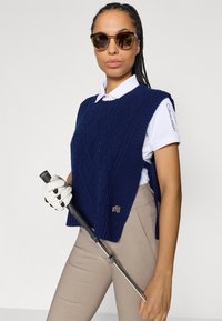 Tengerészkék kötött mellény texturált gyémántmintával, fehér galléros ing fölött viselve. Bézs nadrág és golfkesztyű; golfütőt tart.