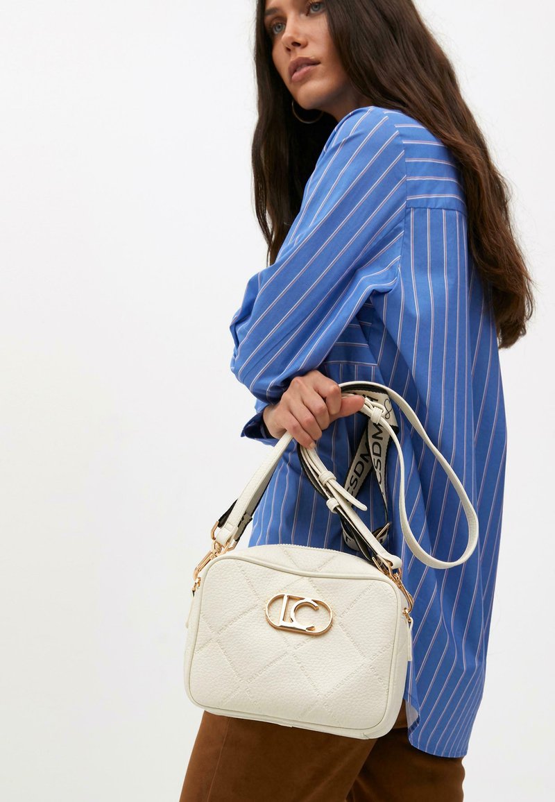 A woman with long, dark hair wears a blue striped shirt and holds a white handbag with a gold logo and patterned finish.