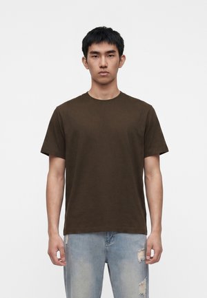 Jeune homme aux cheveux noirs courts, portant un t-shirt uni marron foncé et un jean bleu clair déchiré, debout devant un fond blanc.