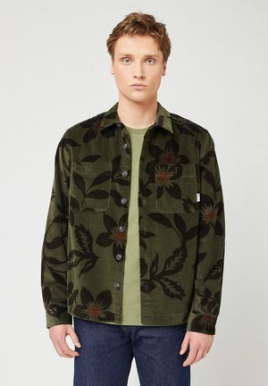 Groene corduroy overhemd met een groot donker bloemenpatroon, met knopen aan de voorkant, twee borstzakken en een klassieke kraag, gedragen over een groene t-shirt.