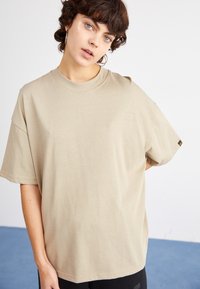 Beige bavlněné tričko s krátkým rukávem, kulatým výstřihem a malým embosovaným logem na přední straně. Měkká textura a volný střih.