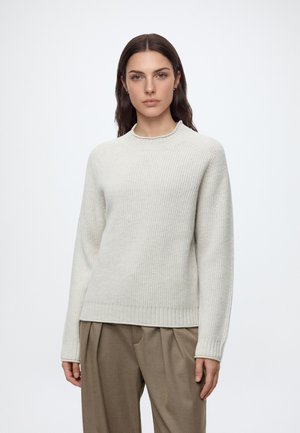 Femme aux cheveux raides et foncés portant un pull léger en maille et un pantalon marron ample, debout sur un fond blanc.