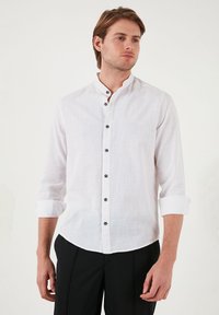 Camisa de lino blanca con cuello mandarín, mangas largas arremangadas en los puños, cierre de botones con botones oscuros y un acabado texturizado.