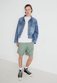 Jachetă din denim de culoare bleu deschis, cu închidere cu nasturi, tricou alb, pantaloni scurți verzi și adidași negri cu gât înalt, cu șireturi albe, plasată pe un fundal simplu.