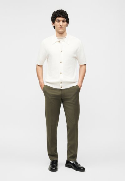 Άνδρας που στέκεται με τα χέρια στις τσέπες φορώντας λευκό πουκάμισο με κουμπιά και κοντά μανίκια, ελαι πράσινο παντελόνι και μαύρα μοκασίνια σε λευκό φόντο.