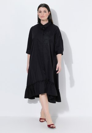 Donna con capelli lunghi e scuri che indossa un abito midi nero e ampio con orlo arricciato e tacchi rossi aperti, in piedi su sfondo bianco.